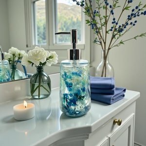 May include: A clear liquid soap dispenser filled with blue and white dried flowers. The dispenser has a silver pump and sits on a white countertop next to a lit candle, a vase of white roses, and blue towels. A vase of blue berries is in the background.