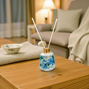 Könnte beinhalten: Ein klarer Glas-Aromatherapie-Diffusor mit blauen floralen Akzenten und zwei Holzstäbchen. Der Diffusor hat einen goldfarbenen Deckel und steht auf einem Holztisch. Eine kleine Schüssel und ein gefaltetes Handtuch befinden sich ebenfalls auf dem Tisch.