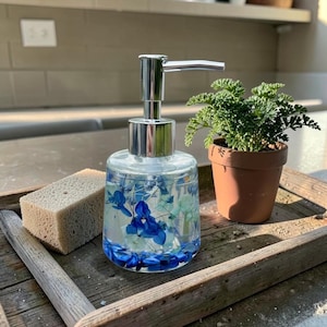 May include: A clear glass soap dispenser with a silver pump, filled with blue and white dried flowers and blue stones. Beside it sits a rectangular beige sponge and a small green plant in a terracotta pot, all on a wooden tray.