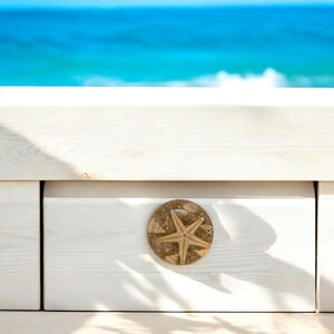 May include: A white wooden drawer with a round, decorative knob featuring a starfish design. The knob is tan and brown, with a starfish in the center. The background shows a bright blue ocean.