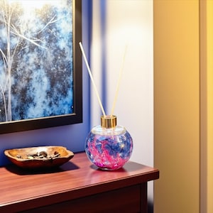 Peut inclure: Un diffuseur décoratif en verre avec un bord doré et deux bâtonnets blancs. La bouteille sphérique est remplie d'éléments décoratifs roses et bleus. Le diffuseur est posé sur une surface en bois foncé, à côté d'un bol en bois et d'un tableau encadré.