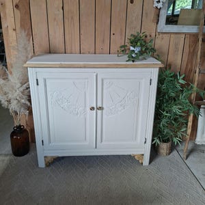 Peut inclure: Une armoire blanche à deux portes dotée d'un plateau en bois naturel et de détails sculptés en forme d'éventail sur chaque panneau de porte. Le meuble repose sur des pieds sculptés décoratifs et possède de petits boutons métalliques ronds.