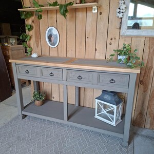 Peut inclure: Une console rectangulaire avec un plateau en bois naturel et une base peinte en gris. Le meuble comporte quatre tiroirs avec des poignées en métal foncé et une étagère inférieure. Une lanterne blanche est posée sur l'étagère et une plante.