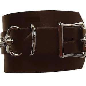 Peut inclure: Un large bracelet manchette en cuir marron foncé avec un fermoir et une quincaillerie en métal argenté. Le bracelet est doté d'une grande boucle et d'un fermoir pivotant pour la fermeture. Le design est simple et audacieux.