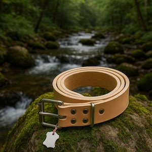 Könnte beinhalten: Ein aufgerollter, hellbrauner Ledergürtel mit einer silberfarbenen Schnalle und Nieten. Der Gürtel wird auf einem moosbedeckten Felsen mit einem verschwommenen Bach und Wald im Hintergrund präsentiert. Ein kleines, silberfarbenes Etikett hängt am Gürtel.