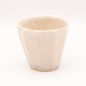 Könnte beinhalten: Eine kleine, cremefarbene Keramiktasse mit facettiertem Design. Die Tasse hat eine leicht konische Form und eine glatte, unglasierte Oberfläche. Der Innenraum ist in einem helleren Beigeton gehalten. Die Tasse ist wahrscheinlich handgefertigt und hat eine rustikale Ästhetik.