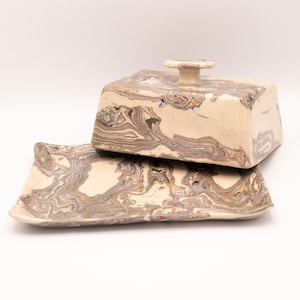 Könnte beinhalten: Ein Keramik-Butterdose mit Deckel, cremefarben und einem marmorierten Muster in Braun-, Grau- und Blautönen. Der Deckel hat einen kleinen, runden Griff. Die Dose steht auf einer passenden rechteckigen Basis.