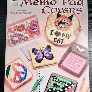 May include: A craft instruction booklet titled Annie's Attic Memo Pad Covers by Glenda Chamberlain. The cover displays various plastic canvas projects including a floral design, a cat face, a butterfly, a peace sign, and a shoe graphic.