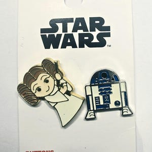 May include: Two Star Wars enamel pins on a white backing card. One pin depicts Princess Leia with brown hair buns and a white dress. The other pin is the droid R2-D2 in white and blue. The card features the Star Wars logo and the word Disney.