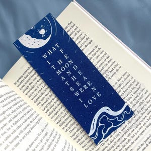 May include: A navy blue bookmark with white text that reads "WHAT I THE MOON AND THE SEA WERE I LOVE." The bookmark features a moon and wave design. The bookmark is placed in an open book.
