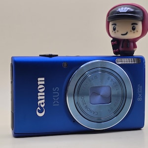 Pode incluir: Uma câmara digital Canon IXUS azul com acabamento metálico, apresentando uma lente circular com o texto 8x optical zoom. Uma pequena figura de uma personagem com um capuz bordô e um boné preto está posicionada no canto superior direito.