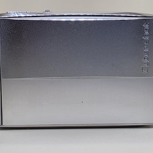 Pode incluir: Uma câmara digital Sony Cyber-shot DSC-T5 prateada com corpo retangular. A parte frontal apresenta um acabamento metálico com o logótipo da Sony no lado esquerdo, o nome do modelo DSC-T5 e a marca Cyber-shot impressa no lado direito.