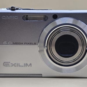May include: Silver Casio Exilim digital camera with a zoom lens. The camera has a 6.0 megapixel resolution and a built-in flash. The camera has a wrist strap attached.