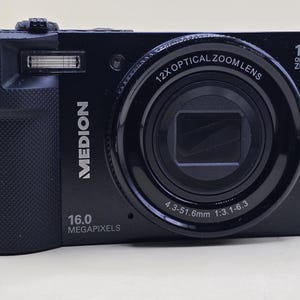 Puede incluir: Una cámara digital negra con lente de zoom óptico 12x, 16.0 megapíxeles y la marca MEDION impresa en el frente. El barril del objetivo muestra el texto 12x OPTICAL ZOOM LENS y 4.3-51.6mm 1:3.1-6.3, con una unidad de flash integrada.