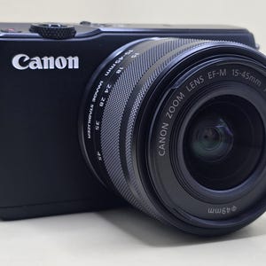Puede incluir: Una cámara digital Canon negra con un objetivo Canon Zoom Lens EF-M 15-45mm 1:3.5-6.3 IS STM. El barril del objetivo muestra marcas de distancia focal de 15 a 45mm y un diámetro de filtro de 49mm. El cuerpo tiene una empuñadura texturizada y un logo Canon.