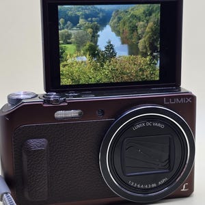 May include: A dark brown Lumix digital camera with a flip-up screen displaying a landscape photo of a river winding through a forest. The lens barrel is labelled Lumix DC Vario 1:3.3-6.4/4.3-86 ASPH. A grey wrist strap is attached to the side.