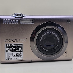 Op de afbeelding: Een metallic bronskleurige Nikon Coolpix digitale camera met een 4x groothoek optische zoom Nikkor-lens, een 12,0 megapixel sensor en een 7,5 cm touchscreen. De voorzijde toont de merknaam, het model en de technische specificaties.