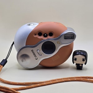 Könnte beinhalten: Eine runde, pfirsichfarbene Canon Ixus Concept Kamera mit weißem Oberteil, silbernem Objektivgehäuse und eingebautem Blitz. Die Kamera verfügt über einen Ein-Aus-Schalter, ein Objektiv und ein langes, orangefarbenes, gesprenkeltes Seil-Trageband.