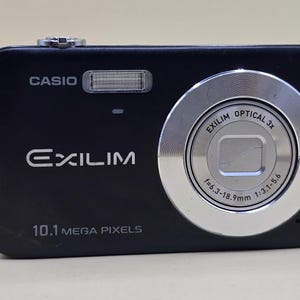 May include: Black Casio Exilim digital camera with a silver lens ring. The camera has a built-in flash and the text "EXILIM" and "10.1 MEGA PIXELS" are visible. The lens has the text "OPTICAL 3x" and "f=6.3-18.9mm".
