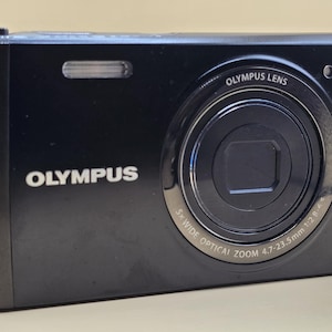 Könnte beinhalten: Eine schwarze Olympus Digitalkamera mit einem silbernen Objektiv. Das Objektiv trägt den Text "Olympus Lens 5x Wide Optical Zoom 4.7-23.5mm 1:2.8-4.4".