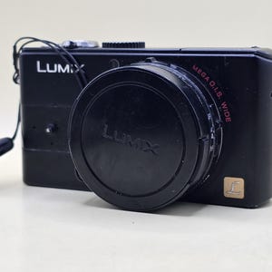 Op de afbeelding: Een zwarte Lumix digitale camera met een opvallende lens en een zwarte lensdop. De camerabehuizing toont de merknaam Lumix in witte letters, een rood Mega O.I.S. Wide label en een klein goudkleurig vierkant logo met een L-embleem.