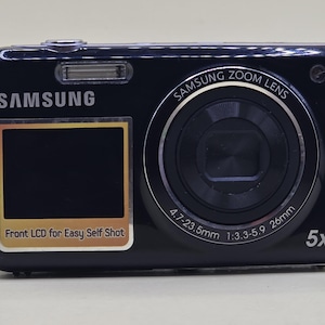 Op de afbeelding: Een zwarte Samsung digitale camera met een LCD-scherm aan de voorzijde met de tekst 'Front LCD for Easy Self Shot' eronder. De lens is gelabeld met 'SAMSUNG ZOOM LENS 4.7-23.5mm 1:3.3-5.9 26mm' en bevat een 5x optische zoomindicator.