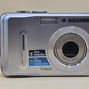 May include: Silver Samsung D860 digital camera with a zoom lens. Features include face detection and digital image stabilisation. The camera has a built-in flash and a wrist strap.