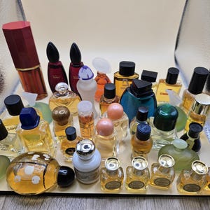 Peut inclure: Une collection de flacons de parfum miniatures de différentes formes et couleurs. L'assortiment comprend des flacons avec des bouchons noirs, bleus et dorés, ainsi qu'un grand tube de rouge à lèvres bordeaux. Les flacons sont disposés sur une surface claire.