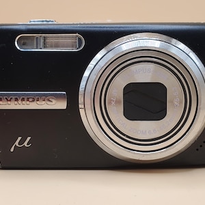 May include: Black Olympus digital camera with a silver lens and flash. The camera features the Olympus logo and the Greek letter mu. The lens has the text "DIGITAL ZOOM 6.6-19.2mm" and "1:3.6-5.3".