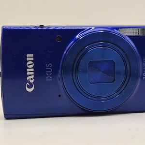 Op de afbeelding: Een koningsblauwe Canon IXUS digitale camera met een 10x optische zoomlens, een ingebouwde flitser en een zwart polsbandje aan de zijkant. De voorkant van de camera toont de merknamen Canon en IXUS in witte letters.