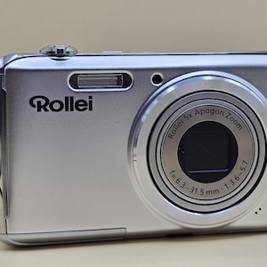 Pode incluir: Câmera digital Rollei prateada com lente zoom. A câmera tem o logotipo Rollei e o texto "Rollei 5x Apogon Zoom" na lente. A lente tem uma distância focal de f=6,3-31,5 mm e uma abertura de 1:3,6-5,7.