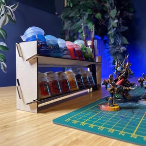 Peut inclure: Un porte-pots de peinture en bois à deux étages, contenant des rangées de petits flacons de peinture. Les flacons sont étiquetés "Citadel Colour" et présentent diverses couleurs, notamment le bleu, le rouge et le jaune. Le porte-pots est posé sur une table en bois avec un tapis de découpe vert.