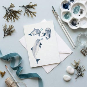 Op de afbeelding: Waterverf wenskaart met drie dolfijnen die in de oceaan zwemmen. De kaart ligt op een witte envelop, omringd door kunstbenodigdheden, waaronder penselen, een palet en blauw lint. Andere elementen zijn gedroogd zeewier en kleine witte stenen.