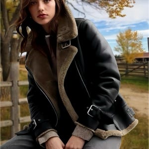 May include: A black leather jacket with a shearling collar and lining. The jacket has silver buckles and a zip closure. The model wears a grey sweater and trousers. The background shows a rural scene with trees and a fence.