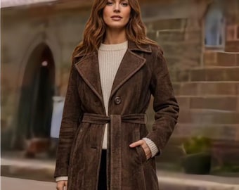 May include: A brown suede coat with a belted waist and a notched collar. The coat has long sleeves and pockets. The model is wearing a cream-colored sweater and black pants. The coat is knee-length.