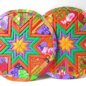May include: Two round fabric pot holders with a starburst design in shades of orange, green, purple, and yellow. The starburst design is made of multiple fabric squares sewn together.