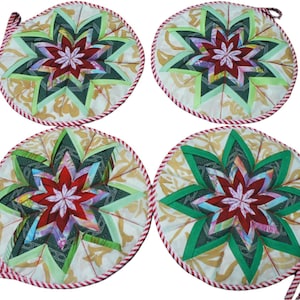 Puede incluir: Cuatro posavasos redondos acolchados con un diseño de estrella en forma de estallido en tonos rojos, verdes y blancos. Los posavasos tienen un fondo blanco con un patrón floral dorado y un borde de rayas rojas y blancas.