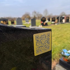 May include: A black granite headstone with a yellow QR code sticker that reads "Scan here for a story to be told." The headstone is in a cemetery with other headstones and green grass in the background.