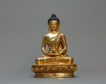 May include: A gold Buddha statue in a seated meditation pose. The statue has a serene expression, with a black bun and intricate detailing on the robes. The base is adorned with a detailed floral pattern. The statue is set against a grey background.