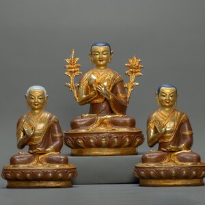 Peut inclure: Trois statues de Bouddha assis, vêtues de robes dorées et marron, avec des accents dorés et des cheveux bleus. Les statues sont sur des socles ornés. La figure centrale tient un élément décoratif doré de chaque côté de la tête.