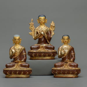 Peut inclure: Trois statues de Bouddha assis, vêtues de robes dorées et marron avec des accents dorés. Les statues sont sur des socles ornés et ont des expressions sereines. Le fond est gris uni.