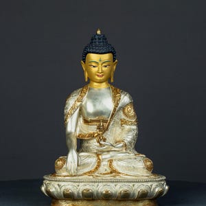 May include: A silver and gold Buddha statue in a seated pose. The statue features a gold face and hands, black hair, and a gold crown. The robe is silver with gold detailing. It rests on a detailed lotus flower base.
