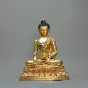 Peut inclure: Une statue dorée de Bouddha en position assise, avec une expression sereine. La statue présente des détails complexes, notamment une coiffure noire, des robes ornées et une base décorative ornée de pierres précieuses colorées. La statue mesure environ 30 cm de haut.