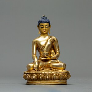 Peut inclure: Une statue dorée de Bouddha en position assise, tenant un petit bol. La statue a une coiffe bleue et des détails dorés. La base est ornée d'un motif floral.