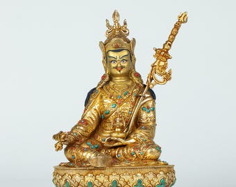 May include: A golden statue of a seated figure, adorned with intricate details and colorful gemstones. The figure wears a crown and holds a decorative staff. The statue is on a detailed base, showcasing a blend of artistry and cultural significance.