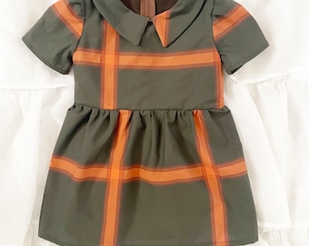 May include: A short-sleeved dress in olive green featuring a bold orange and light orange plaid pattern. The garment has a rounded collar, a gathered waistline, and a brown zipper closure at the back of the neck. The fabric has a smooth finish.