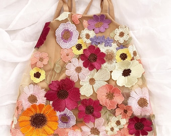 May include: A sleeveless tan dress featuring an overlay of sheer fabric adorned with a dense arrangement of embroidered flowers in shades of orange, magenta, pink, yellow, purple, and cream, with thin shoulder straps tied at the top.