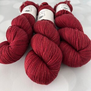 May include: Three skeins of deep red yarn are arranged in a row. Each skein is a plump, rounded shape, with the yarn tightly wound. White labels with a small illustration are visible on top of each skein.