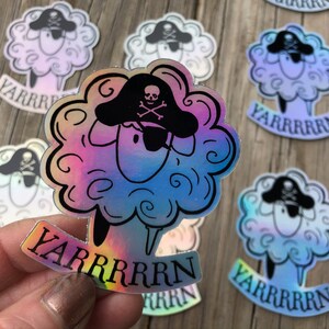May include: A collection of stickers featuring a sheep with a pirate hat and the word "YARRRRRN". The sheep has a fluffy, cloud-like body and a black pirate hat with a skull and crossbones. The stickers have a holographic, rainbow sheen.