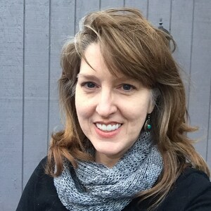 Puede incluir: Una mujer con cabello castaño y una bufanda infinita de punto gris sonríe a la cámara. Lleva una blusa negra y pendientes plateados con detalles turquesa. El fondo es una pared de madera gris.